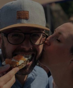 Ein Mann beißt von einem Brot ab und seine Freundin küsst ihn auf die Wange